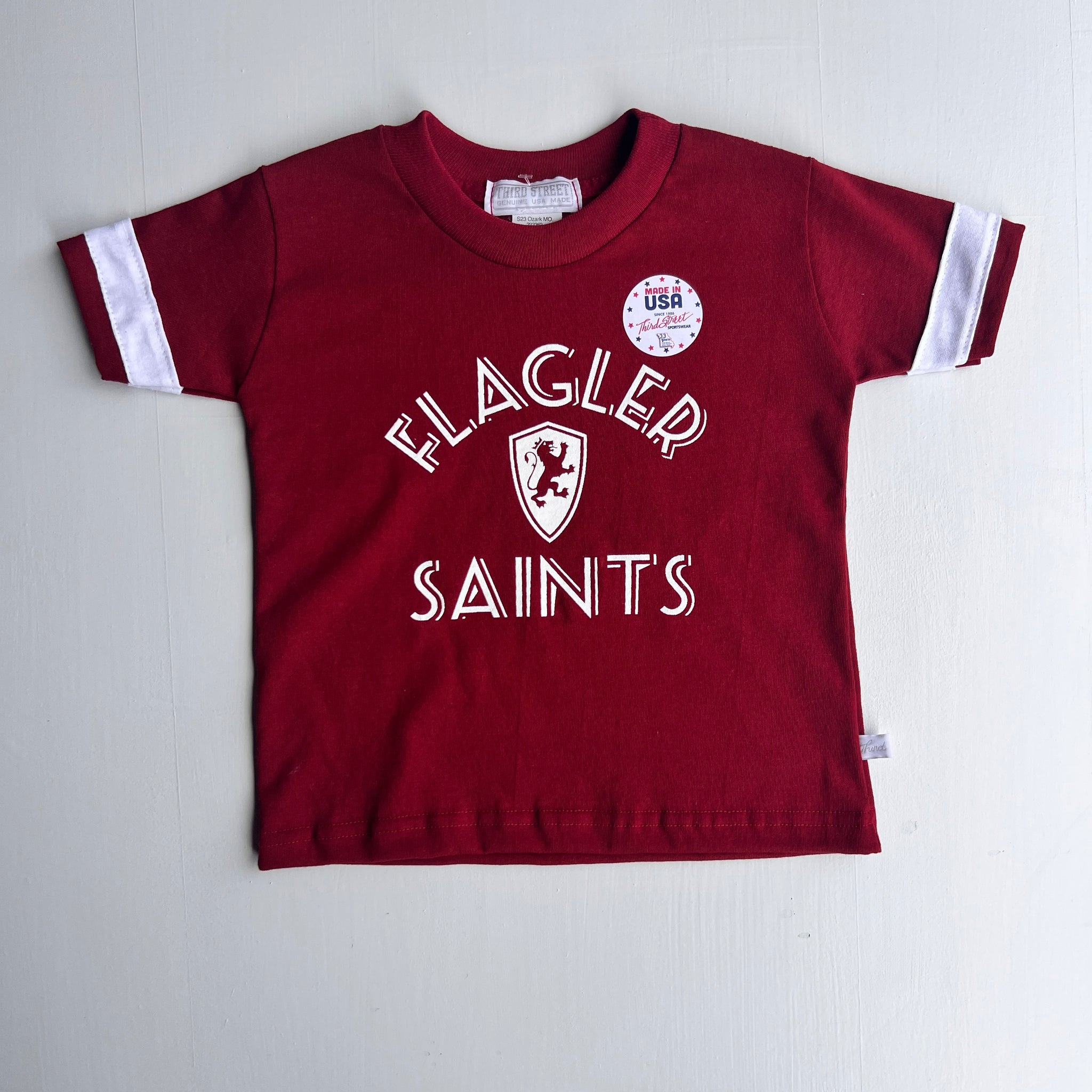 crimson t-shirt with whit stripe on each arm. White imprint saying Flagler over Flagler shield logo over Saints