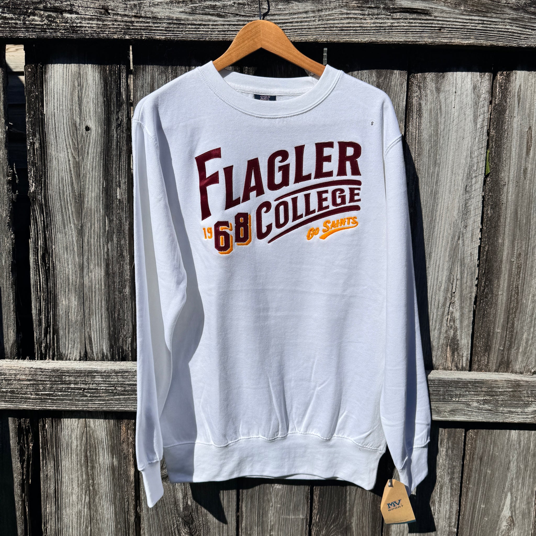 White solid longsleeve crewneck with crimson imprint saying Flagler over swoop over yellow and crimson imprint saying 1968 next to crimson imprint saying College over yellow imprint saying go saints 