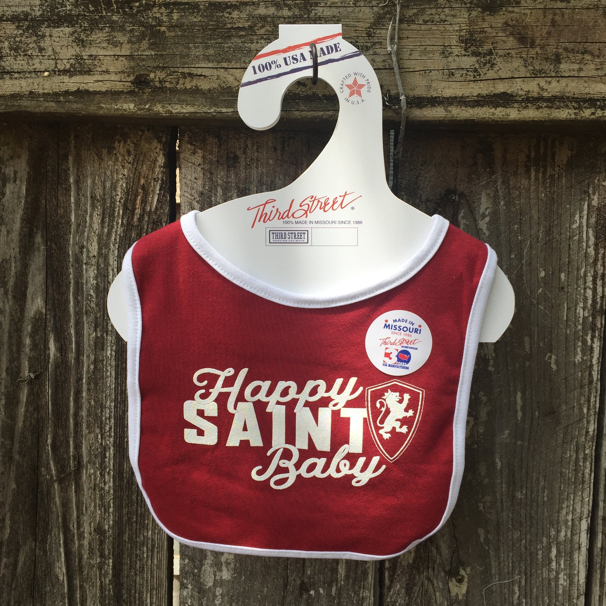 Crimson baby bib with white border. white imprint in center saying Happy over Saint over Baby with white and crimson Flagler College shield logo on left side