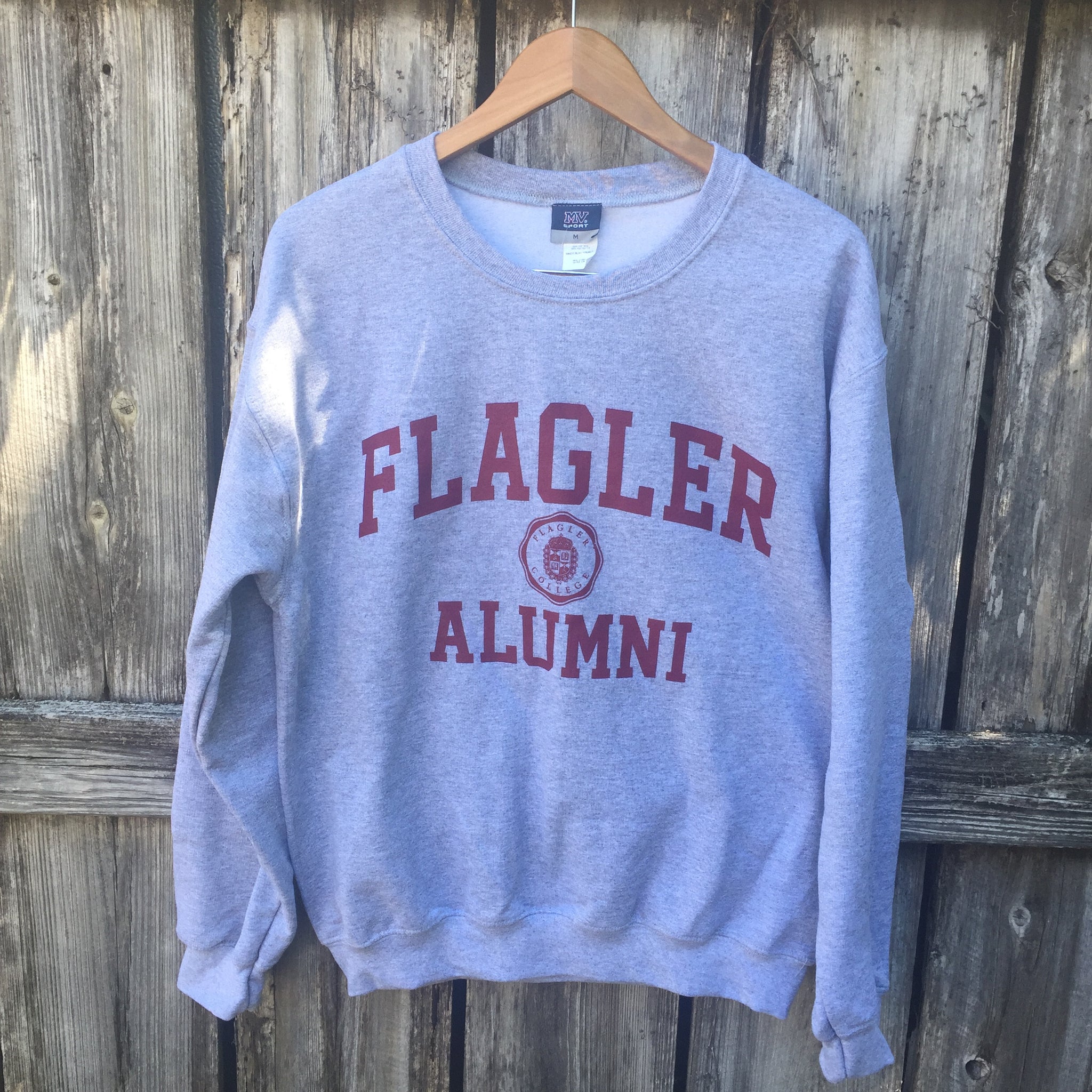 Grey crew neck with Flagler and Alumni printed in red. in between Flagler and alumni is a shield seal also in red