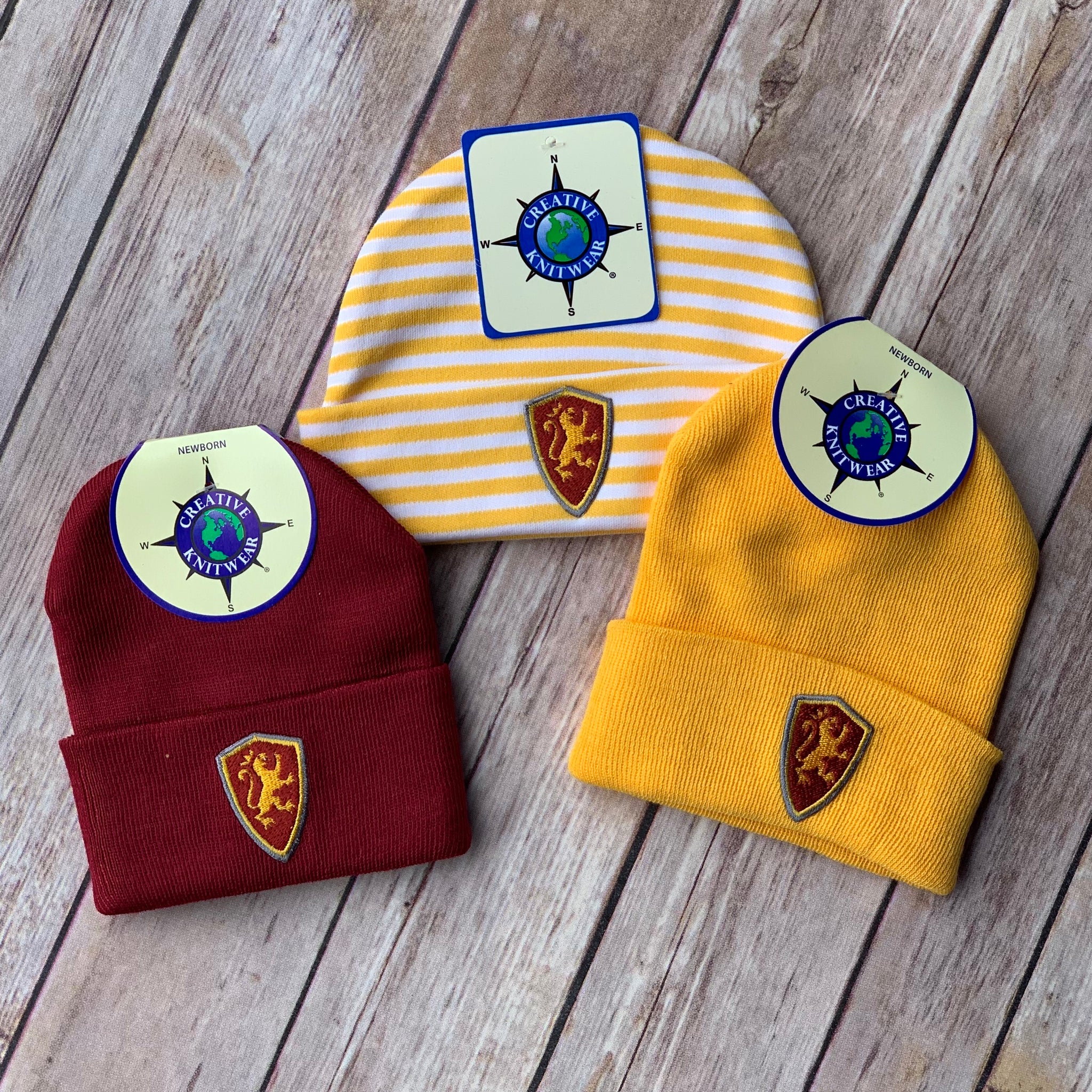 three baby knot caps each with embroidered yellow and red shield patches on the front. the first is solid red, the second has yellow and white stripes and the last is solid yellow